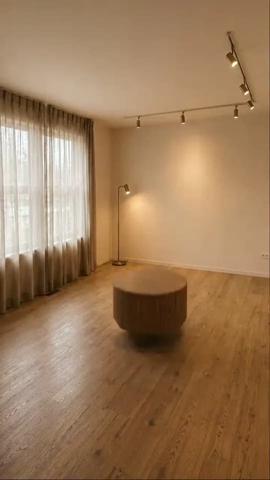 The same room with a slim brass floor lamp by the back wall next to the curtains and a round fluted wooden coffee table hovering slightly above the bare boards in the middle of the floor, its base blurred.