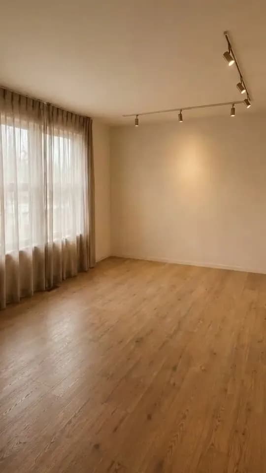The empty room a furniture pop-in reel opens on: full-height sheer linen curtains over a window on the left, a brass ceiling track of spotlights, a warm pool of light on the plain back wall and a bare oak-effect floor.