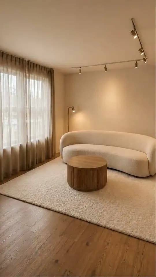 The same room finished, with full-height sheer curtains, a curved cream bouclé sofa, a round fluted wooden table on a cream rug, a floor lamp and lit track lights over oak boards