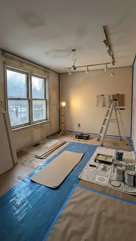 The same room mid-renovation, with the back wall painted beige under a newly fitted track of spotlights, the window wall still bare plaster, blue floor protection, a work light, a stepladder and paint tins on a dust sheet