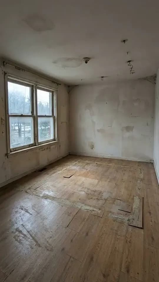 An empty, tired room with a white double window with peeling paint on the left, patched and stained walls, bare wires hanging from the ceiling and worn floorboards with lifted sections