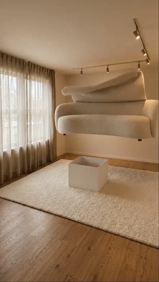 A vertical frame of a room with sheer curtains and a ceiling track of spotlights: an open white box stands on a thick cream rug while a curved cream bouclé sofa floats in the air above and behind it, with a loose fold of the same fabric lying along its top