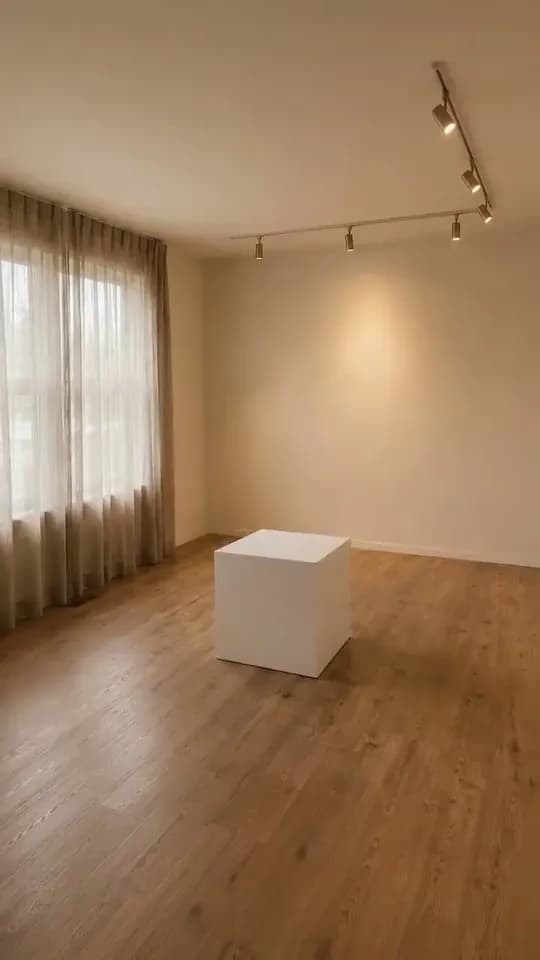 A vertical frame of an empty room with sheer linen curtains over a tall window on the left, a ceiling track of spotlights, warm oak floorboards and a single plain white box standing on the bare floor near the centre